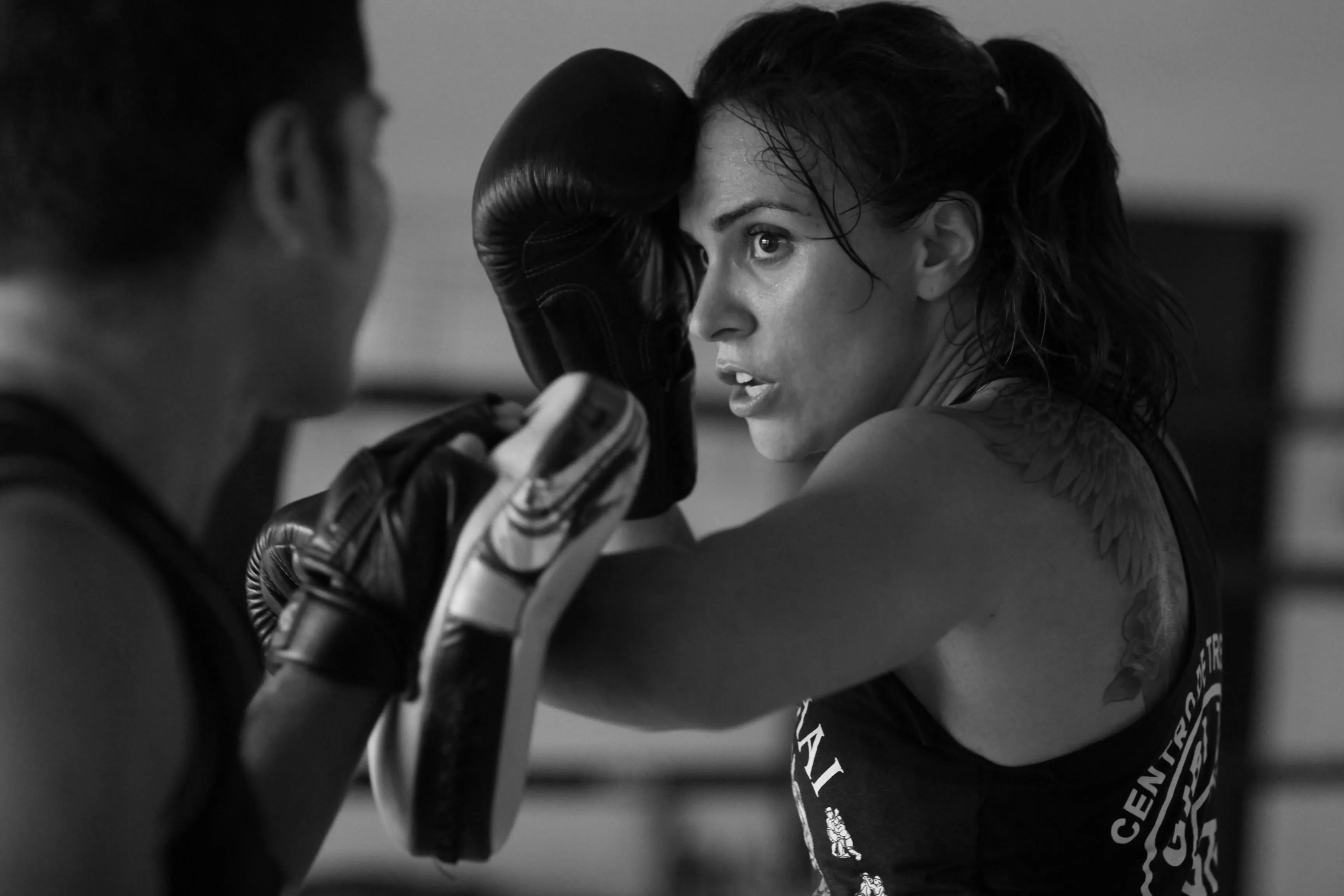 Focused woman with a tattoo trains boxing techniques using yellow and black pads held by a partner.