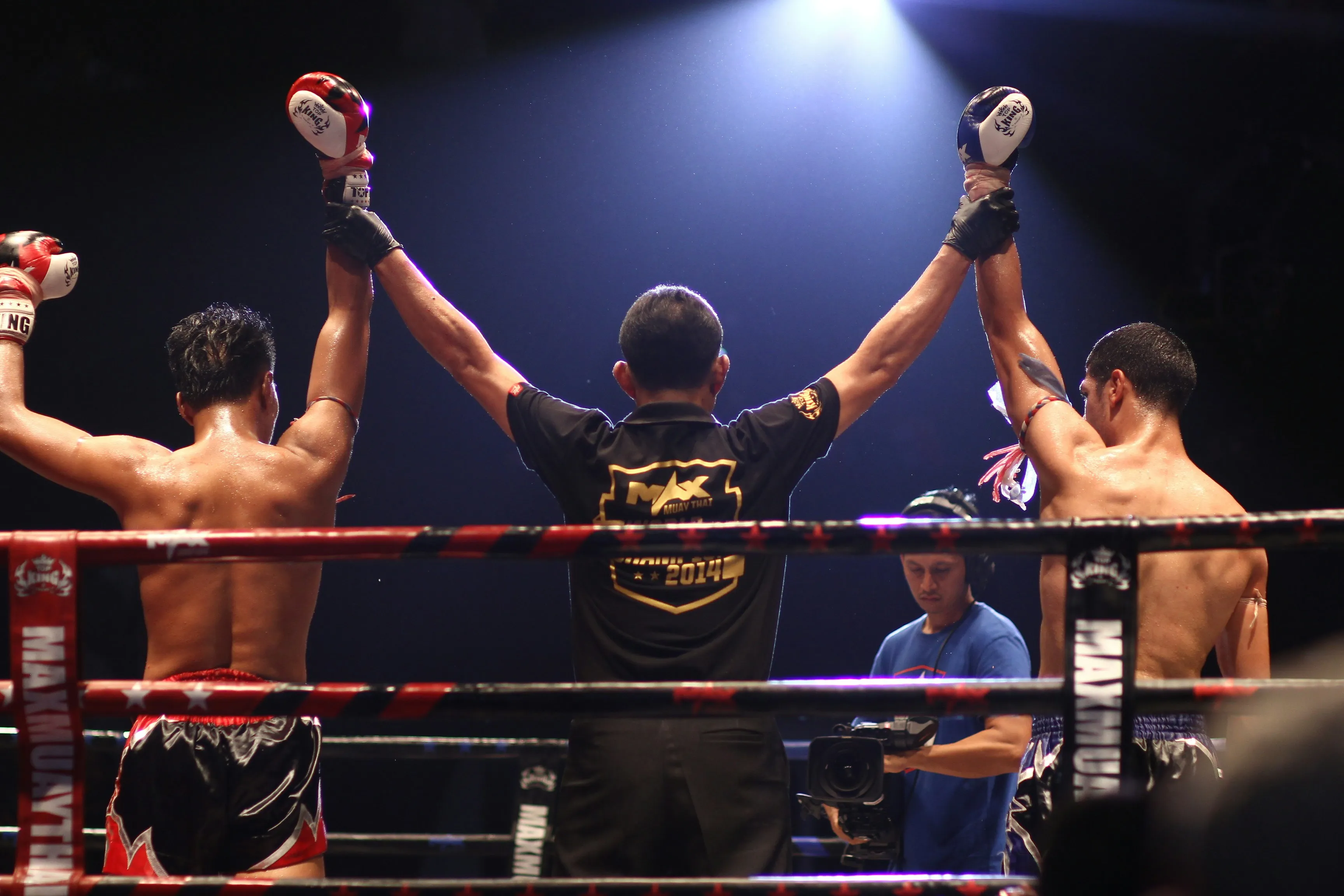 Referee raising the arms of two shirtless Muay Thai fighters in a ring under bright spotlight.