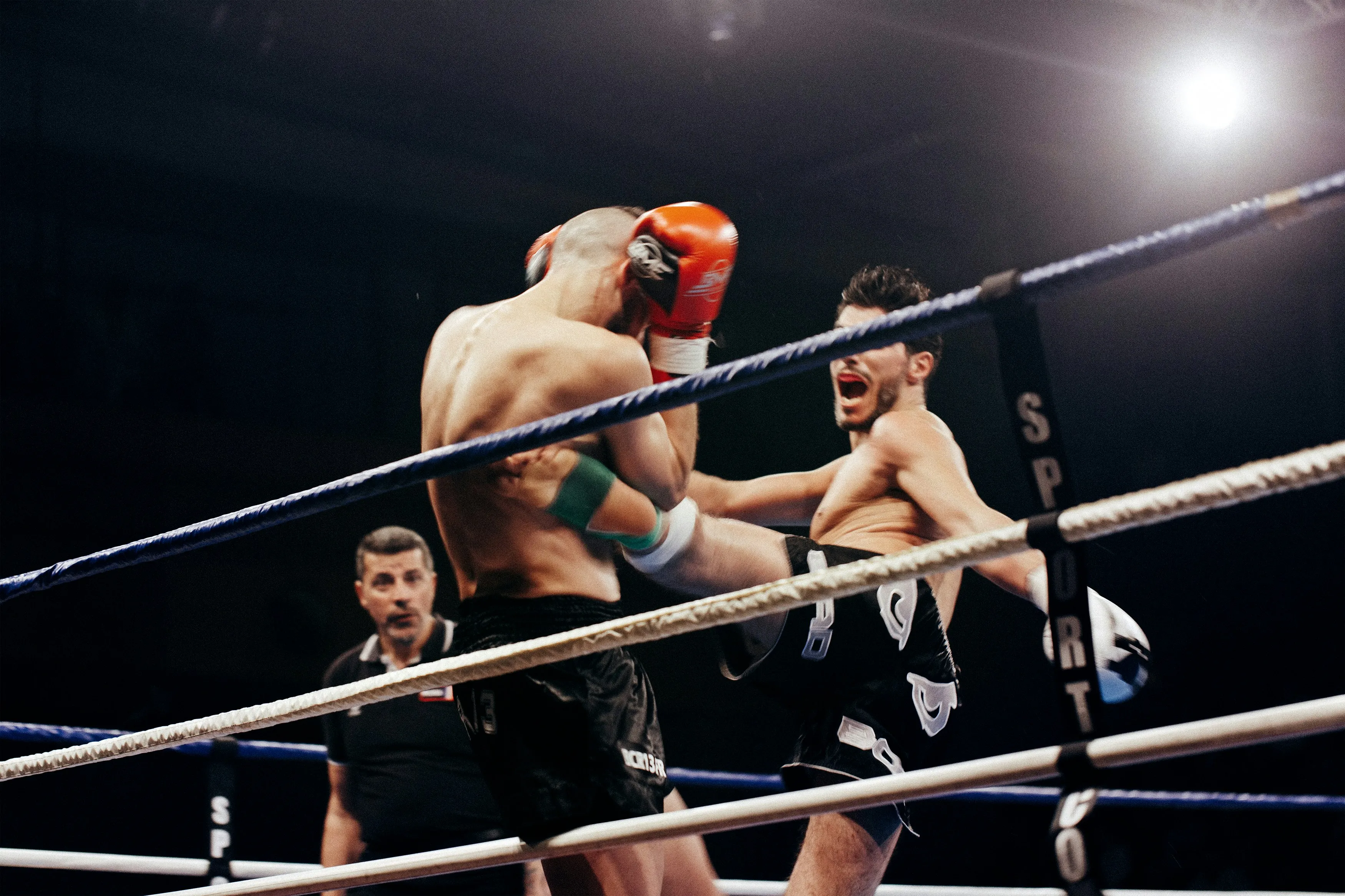 Two male kickboxers in a ring, one delivering a kick to the other's midsection while a referee watches closely.